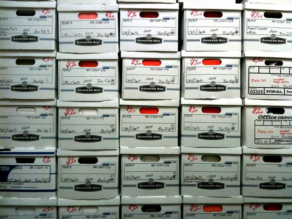 stacked white Bankers Boxes with handwritten labels, organized in rows, labeled with "MERCK", dates, and contents like "CRFs/Charts"