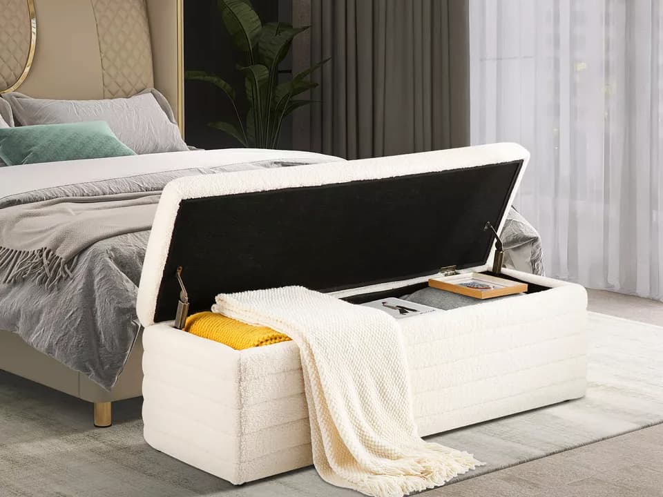 bedroom with upholstered storage bench at foot of bed, bench lid open revealing neatly stored items including yellow and gray blankets, books, and a white knit throw draped over side, modern decor with soft lighting and floor-to-ceiling curtains