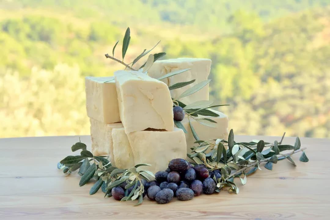 Stacked beige castile soap bars, placed on a wooden surface, surrounded by fresh olives and olive branches, outdoor background with blurred greenery