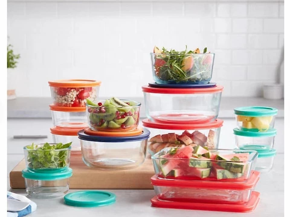 glass food storage containers with colorful lids, filled with cut fruits, vegetables, and greens, arranged on a kitchen counter
