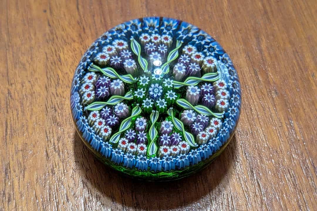 Round glass paperweight, intricate millefiori floral design in green, blue, white, and purple, placed on a wooden surface