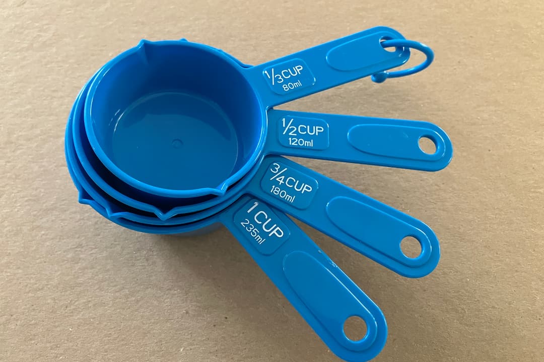 Set of four blue plastic measuring cups labeled 1 cup (235ml), 3/4 cup (180ml), 1/2 cup (120ml), and 1/3 cup (80ml), stacked together on a beige surface