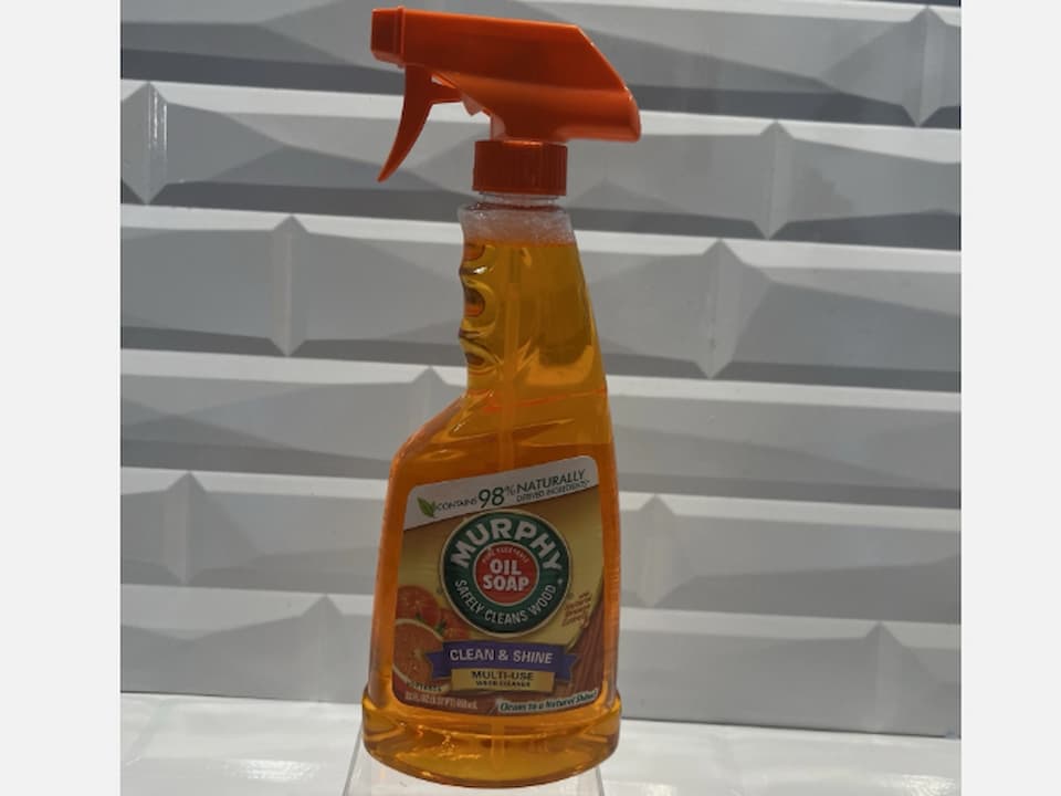 Murphy Oil Soap spray bottle, orange trigger nozzle, amber-colored liquid inside, label reading “Clean & Shine Multi-Use,” placed against a white textured tiled background, slight shadow cast on surface