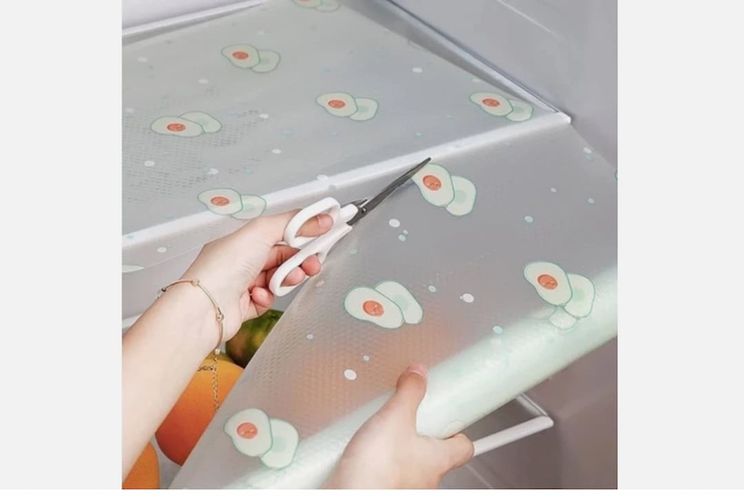 Hands cutting translucent fridge liner with avocado print, using white scissors, fitting liner onto refrigerator shelf