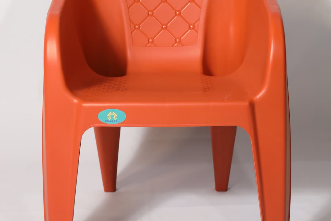 Bright orange plastic chair with armrests and diamond-patterned backrest, brand logo "Prakash" on the front, set against a plain white background