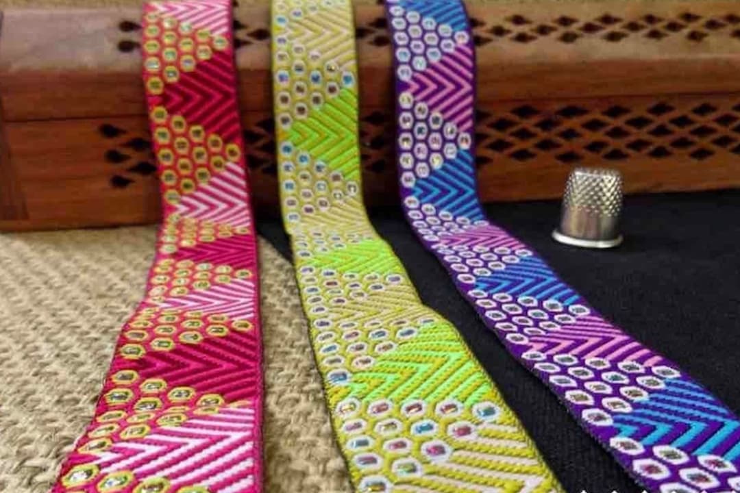 Three colorful woven ribbons with geometric and dotted patterns in red, yellow-green, and blue-purple, placed beside a sewing thimble and a wooden box