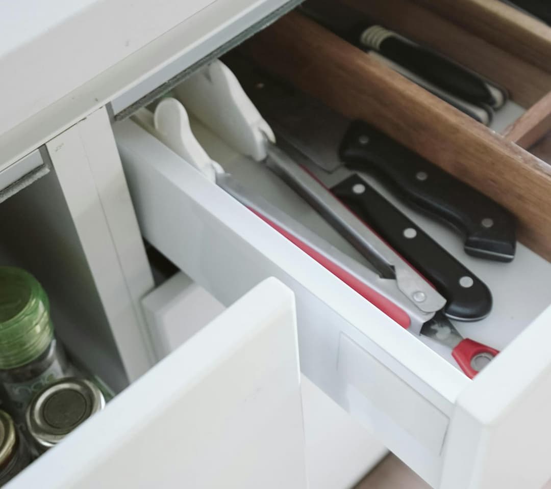 Open white kitchen drawer with wooden dividers organizing knives, tongs, and kitchen tools, spice jars visible in adjacent cabinet