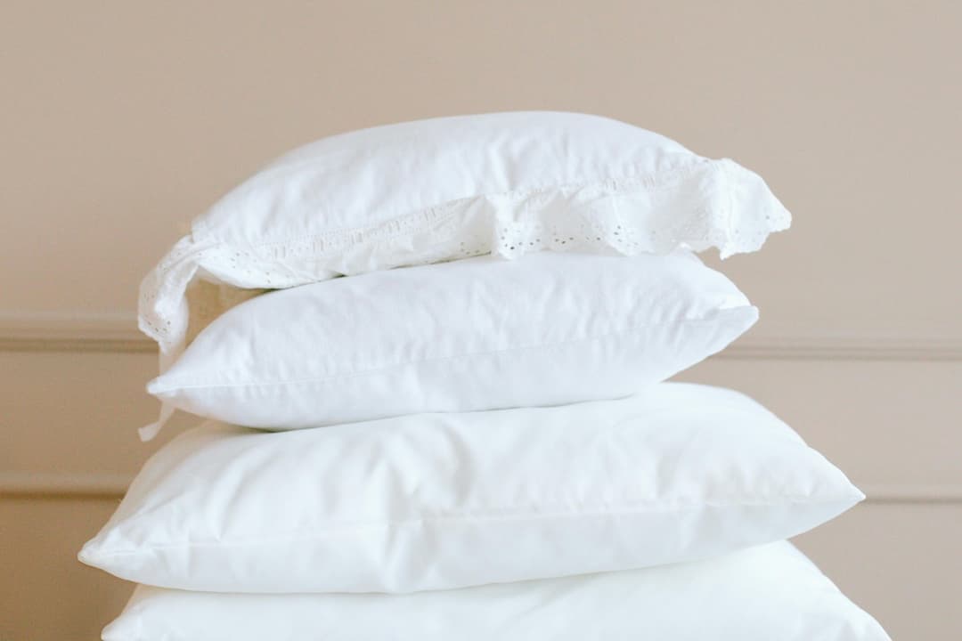 Stack of four white pillows with smooth fabric, the top one featuring a lace trim, placed against a soft beige wall