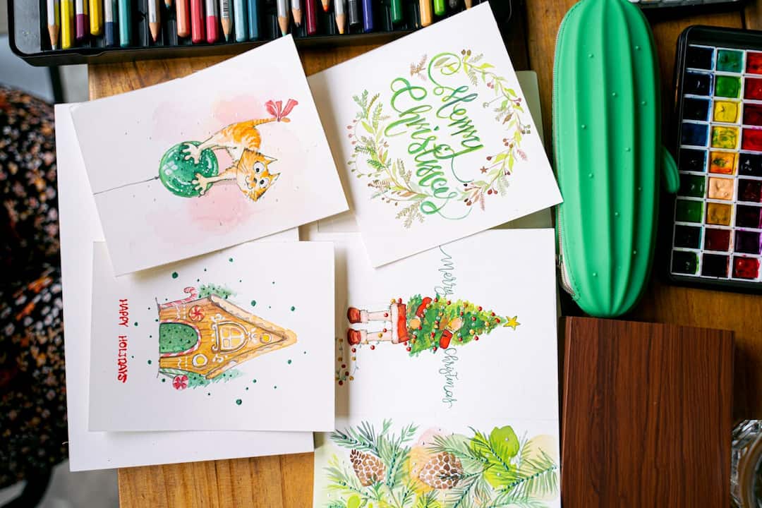 handmade Christmas cards with watercolor illustrations, designs include cat on ornament, gingerbread house, Christmas tree, pine branches, and festive lettering, surrounded by colored pencils, watercolor palette, and green pencil case