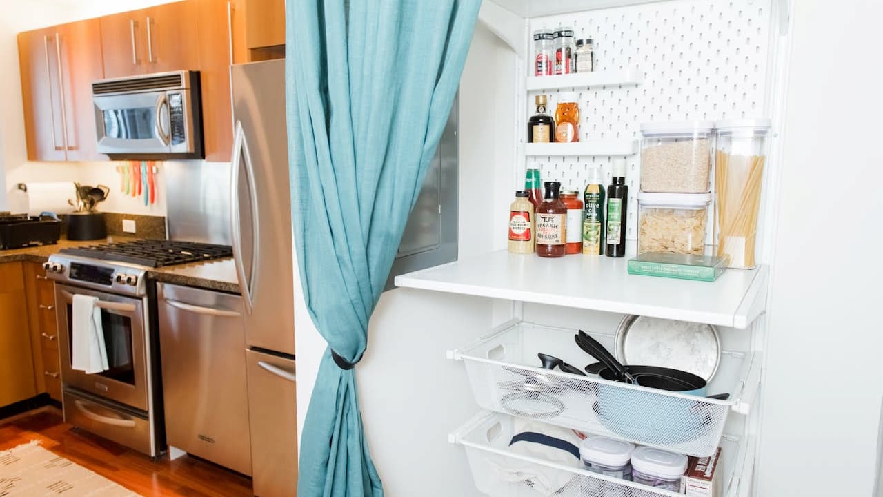 Modern kitchen with stainless steel appliances, open pantry with white shelves, neatly organized spices and dry goods, pots and pans stored below, light blue curtain dividing kitchen and pantry area, warm wooden cabinets