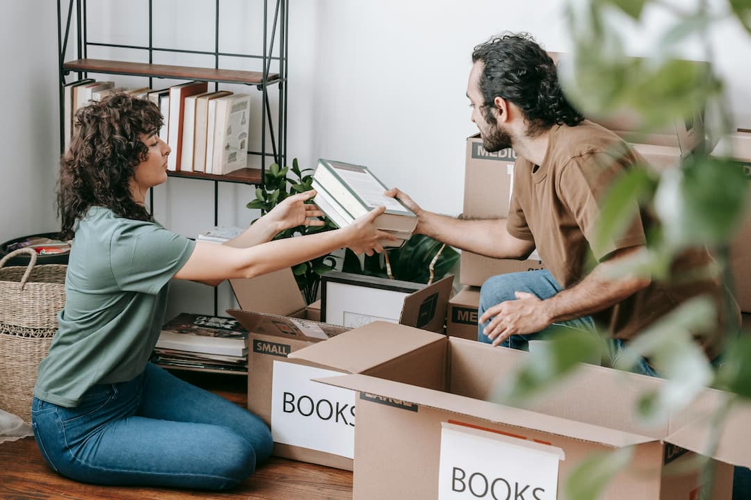 Man and woman packing books into labeled cardboard boxes, surrounded by open boxes and bookshelf in background, mid-move or decluttering