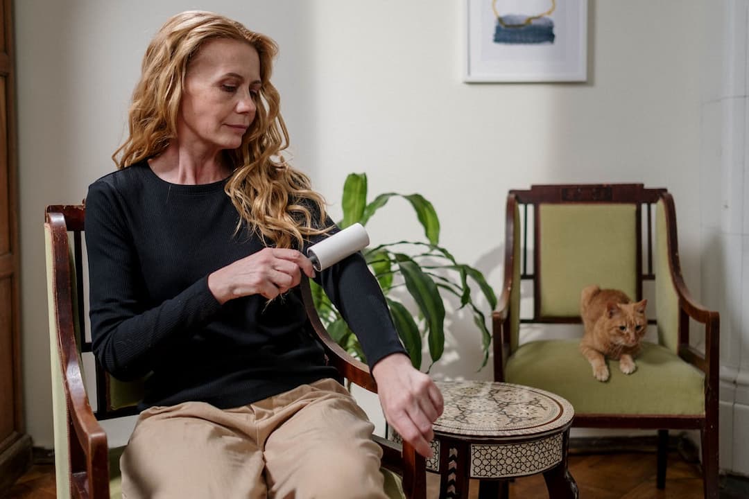Woman sitting on green-upholstered chair, using white lint roller on black sleeve, beige pants, round mosaic side table, orange cat on second chair in background, potted plant and framed artwork on wall