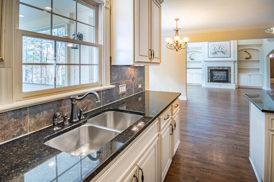 Modern kitchen interior with granite countertops, stainless steel double sink, white cabinets, tiled backsplash, and a view into a living room with hardwood floors, fireplace, and built-in shelves