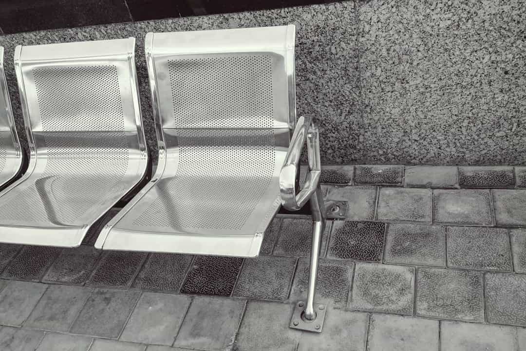 Three silver perforated metal chairs, attached in a row, mounted on a tiled floor, positioned against a speckled stone wall