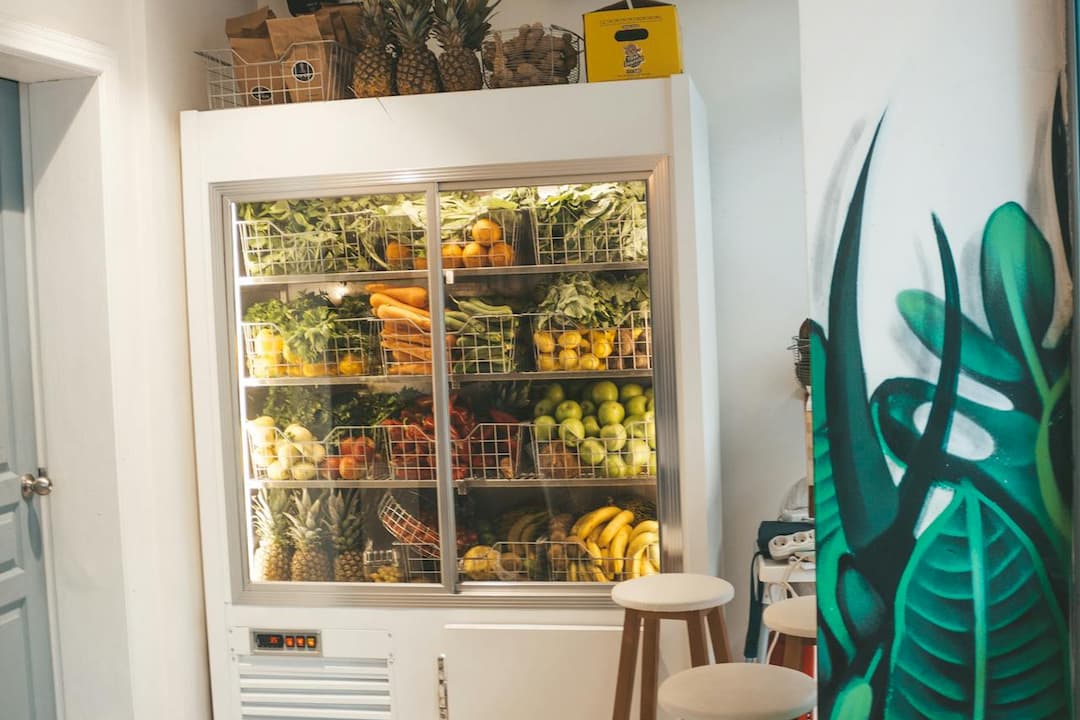 A well-organized refrigerator filled with assorted fresh fruits and vegetables in wire baskets, including pineapples, carrots, bananas, apples, and leafy greens
