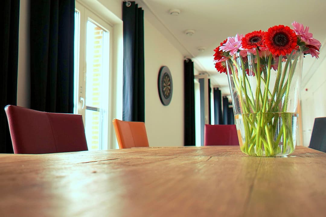 Clear glass vase with red and pink gerbera daisies, placed on a wooden dining table, surrounded by colorful chairs, near large windows with black curtains, bright natural light filling the room