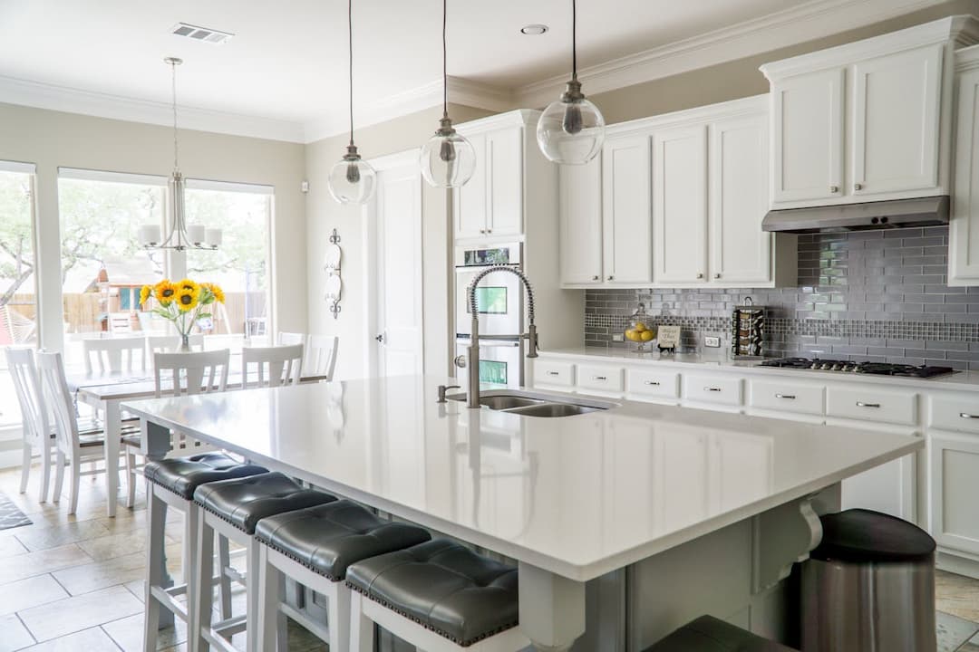 Bright modern kitchen with white cabinets, stainless steel appliances, grey tile backsplash, a large white island with bar stools, and a dining area with a sunflower bouquet centerpiece