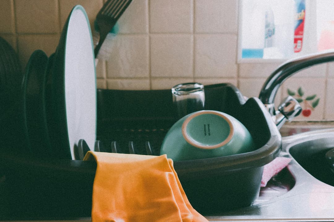 Clean dishes drying in a black dish rack beside a kitchen sink, including plates, a bowl, and a glass, with a yellow rubber glove draped over the edge