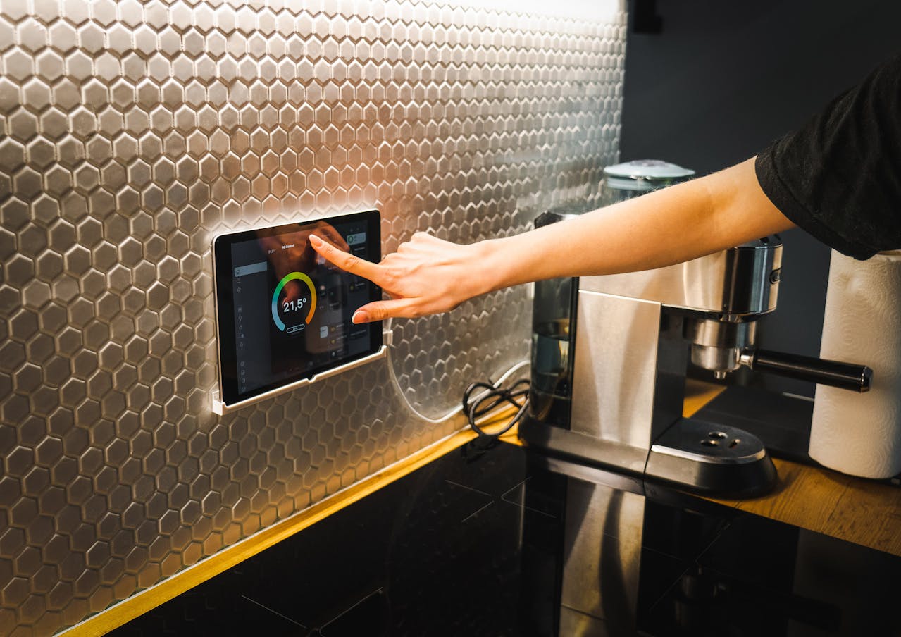 Person adjusting a wall-mounted smart home control tablet, metallic honeycomb backsplash, espresso machine on wooden countertop