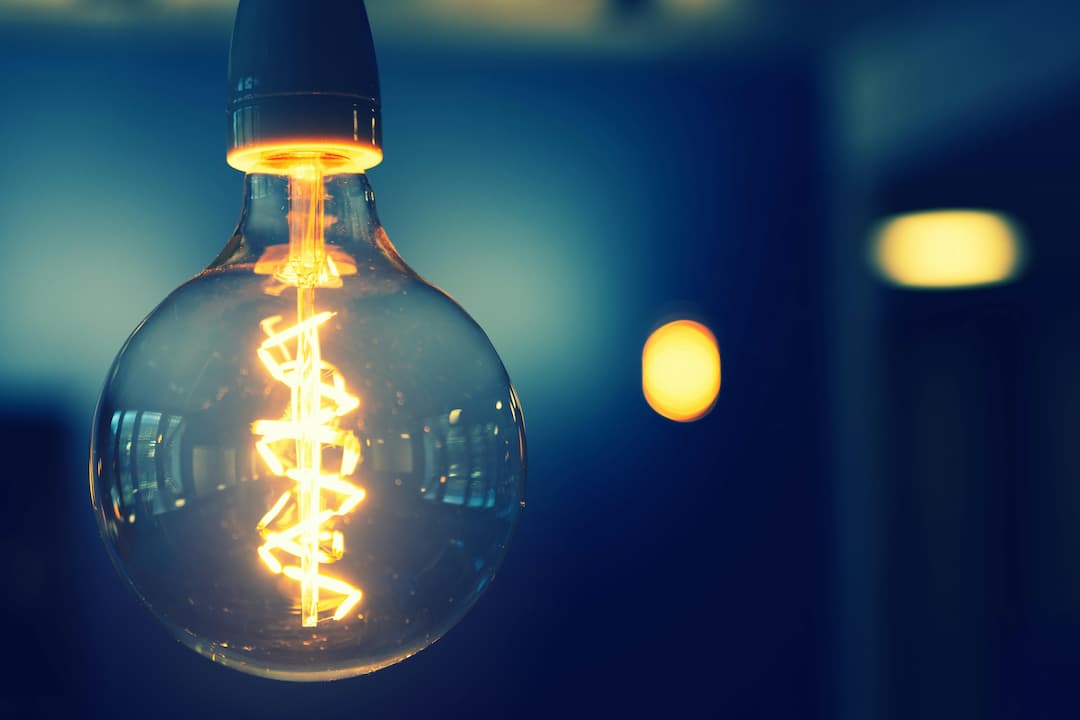 Close-up of a glowing filament light bulb against a blurred dark blue background, with warm yellow bokeh lights in the distance