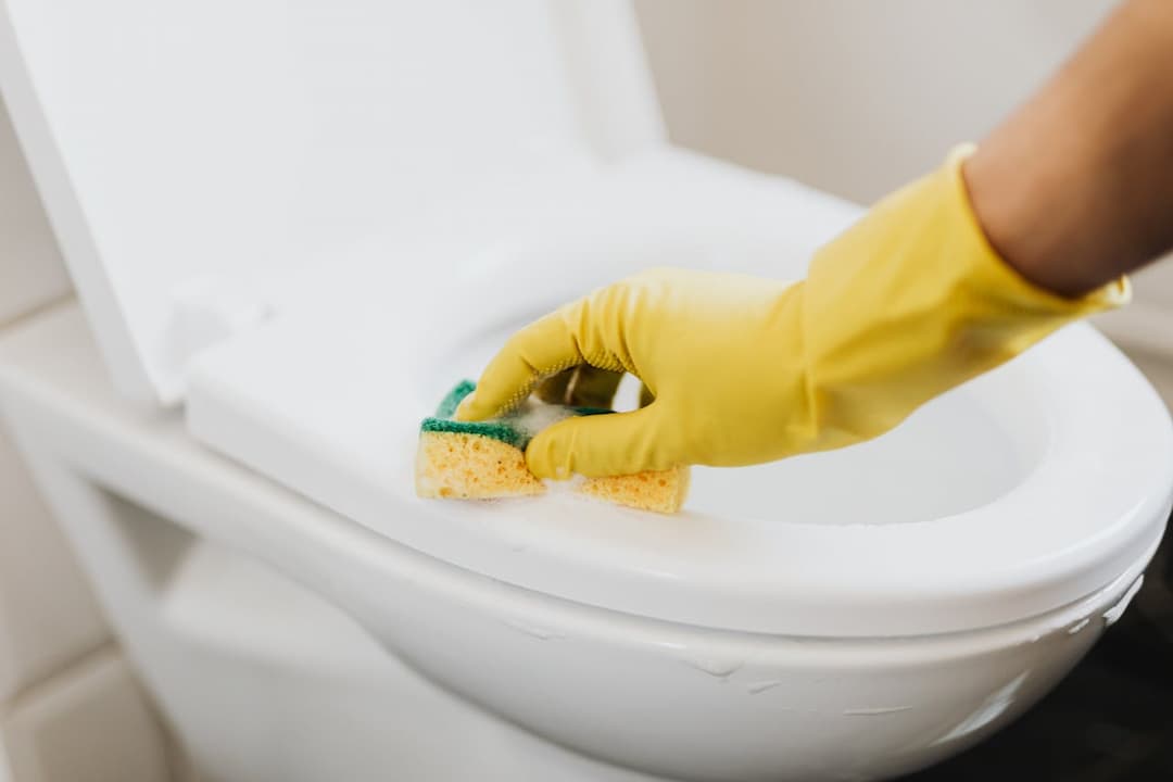 Person wearing yellow gloves scrubbing a white toilet seat with a yellow and green sponge, visible soap suds, toilet lid ope