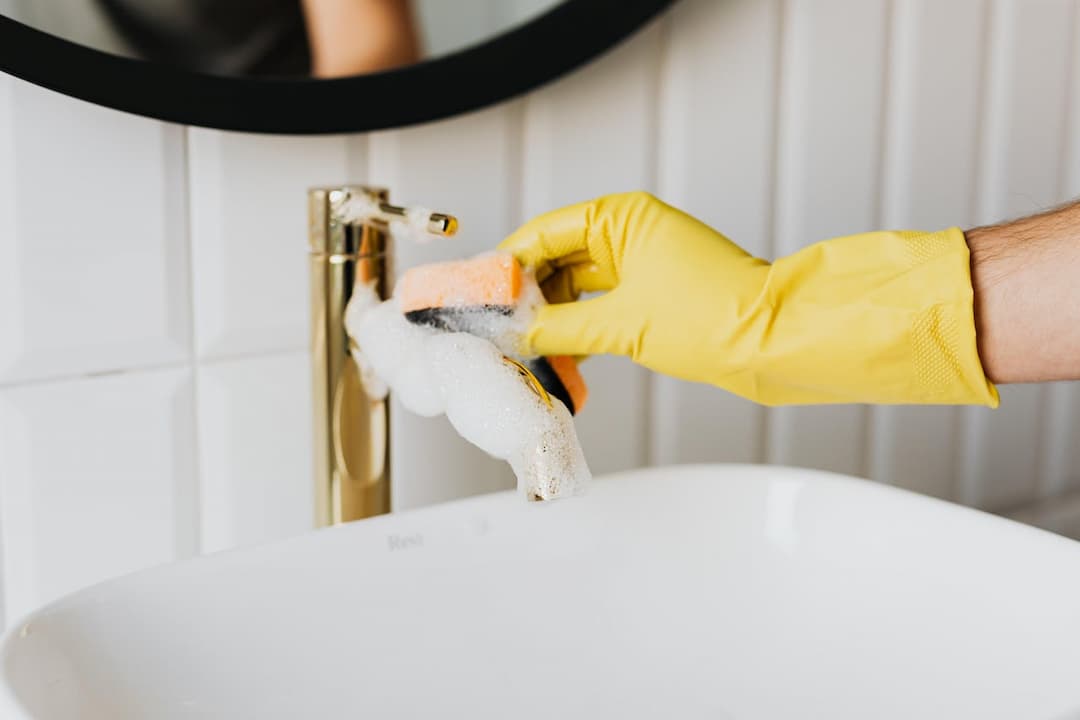 Person wearing yellow gloves scrubbing a gold bathroom faucet with a soapy sponge, white sink below, tiled wall background