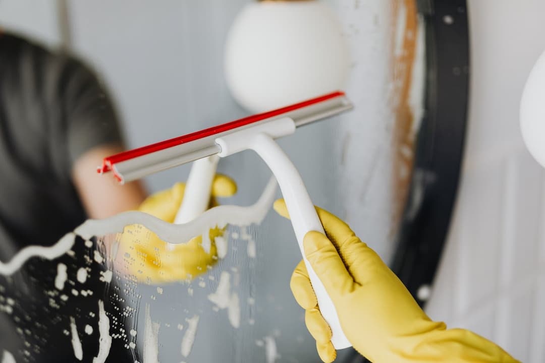 Gloved hands using a white and red squeegee, wiping soap from a mirror, reflection partially visible, cleaning foam along the glass surface, round mirror in background