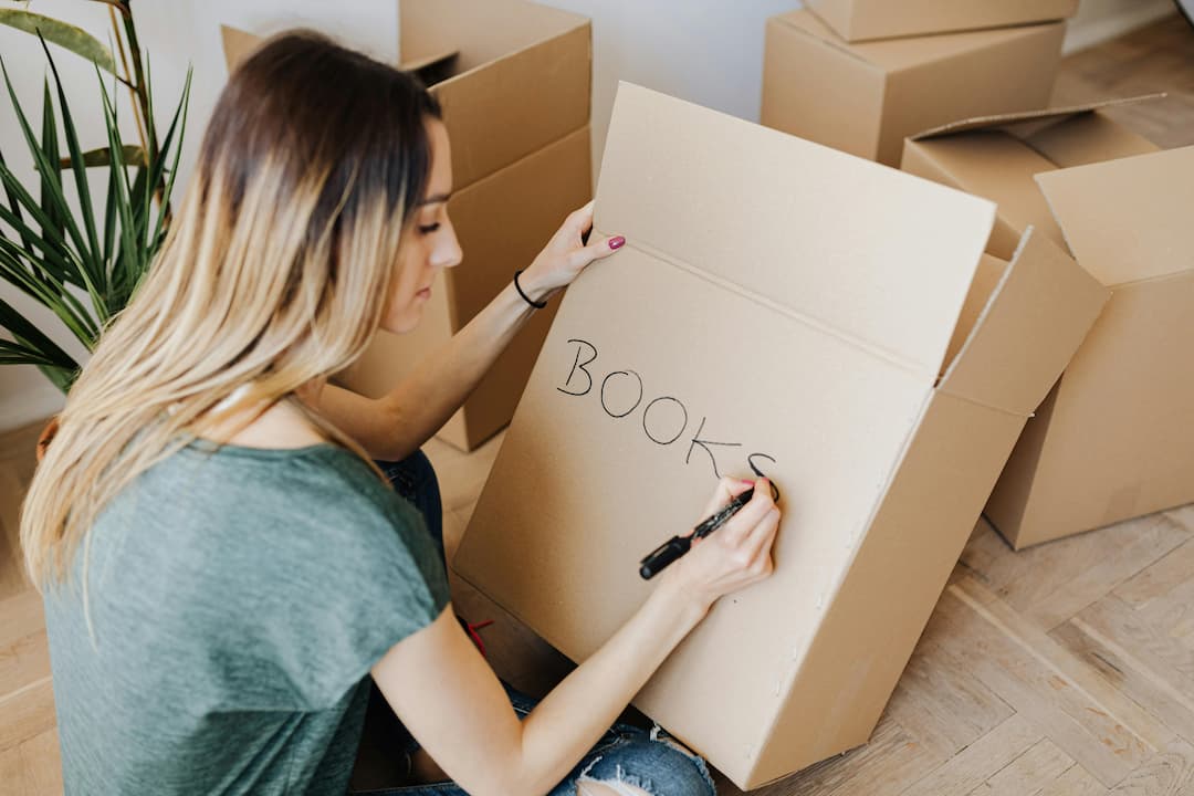 Woman writing "BOOKS" on a cardboard box with a black marker, surrounded by multiple packed and open boxes, seated on wooden floo