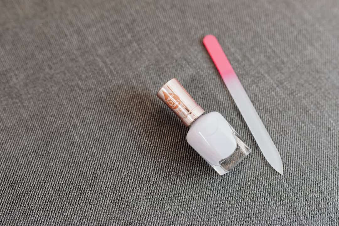 A white nail polish bottle and a glass nail file with a pink tip on a grey fabric surface