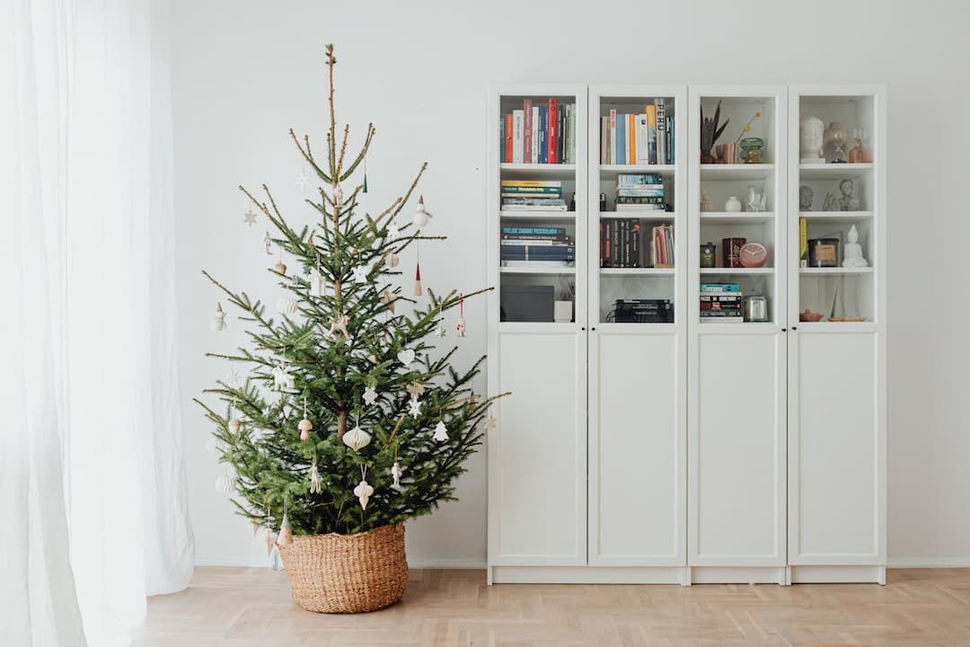 minimalist Christmas tree in woven basket, decorated with neutral ornaments, next to white bookcase with glass doors and books