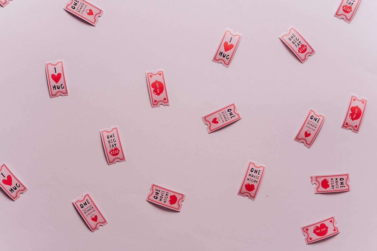 scattered red and pink love-themed tickets on light pink background, printed messages include “1 Hug,” “One Big Fat Kiss,” “One Movie Night,” and “One Dinner Date,” playful and romantic design