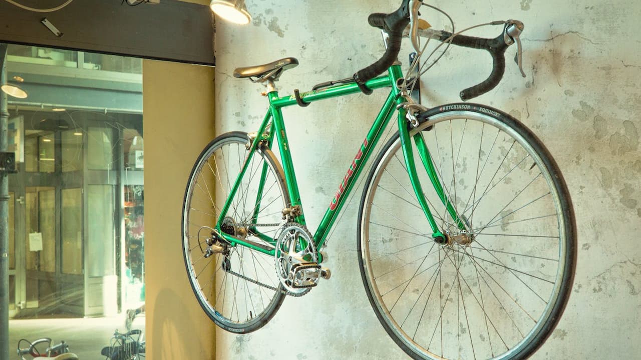 Green road bicycle mounted on a rustic white wall, drop handlebars, thin tires, indoor setting with glass windows and reflections in the background