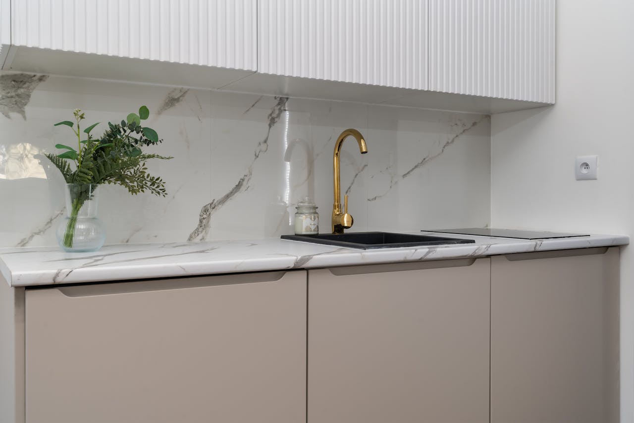 Modern kitchen counter with a gold faucet, black sink, white marble backsplash with gray veining, beige cabinets, and a small glass vase of green foliage