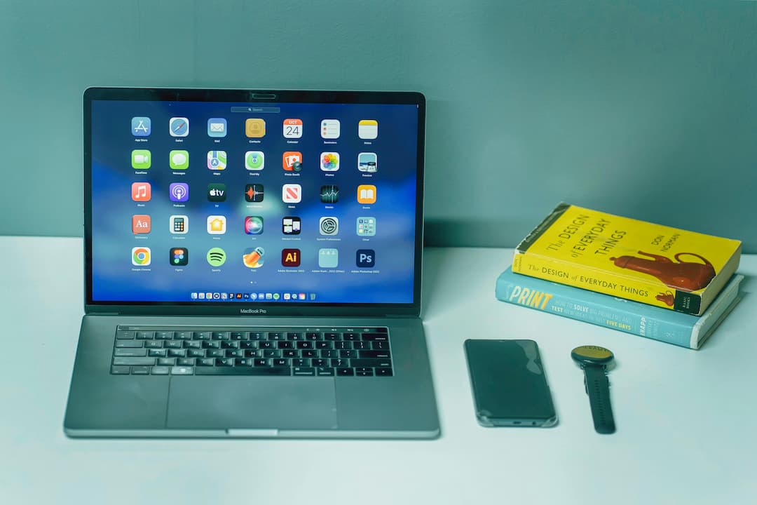 A MacBook Pro displaying application icons on a blue-themed screen, placed on a white desk beside a smartphone, a smartwatch, and two stacked design books 