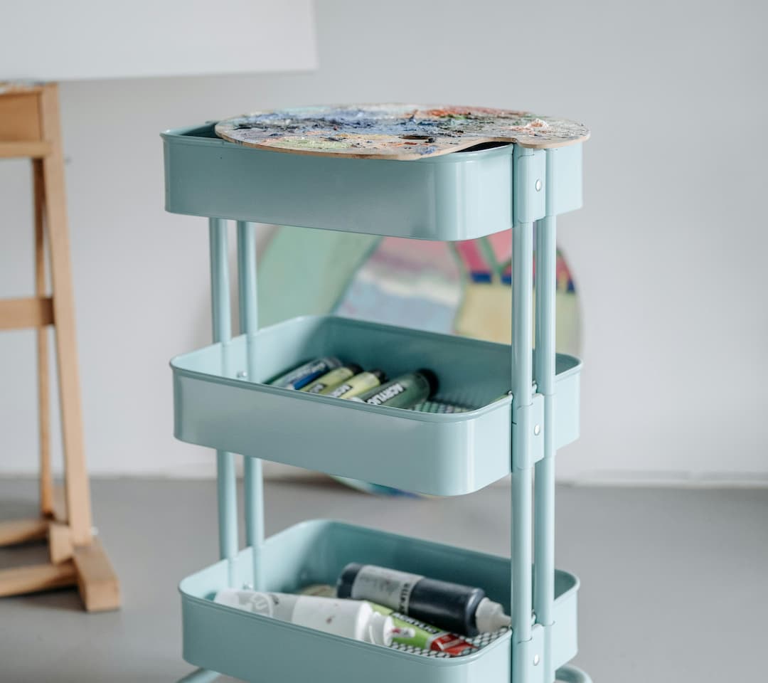 Light blue three-tier rolling cart, top shelf holding a paint palette, middle and bottom shelves containing tubes of acrylic paint
