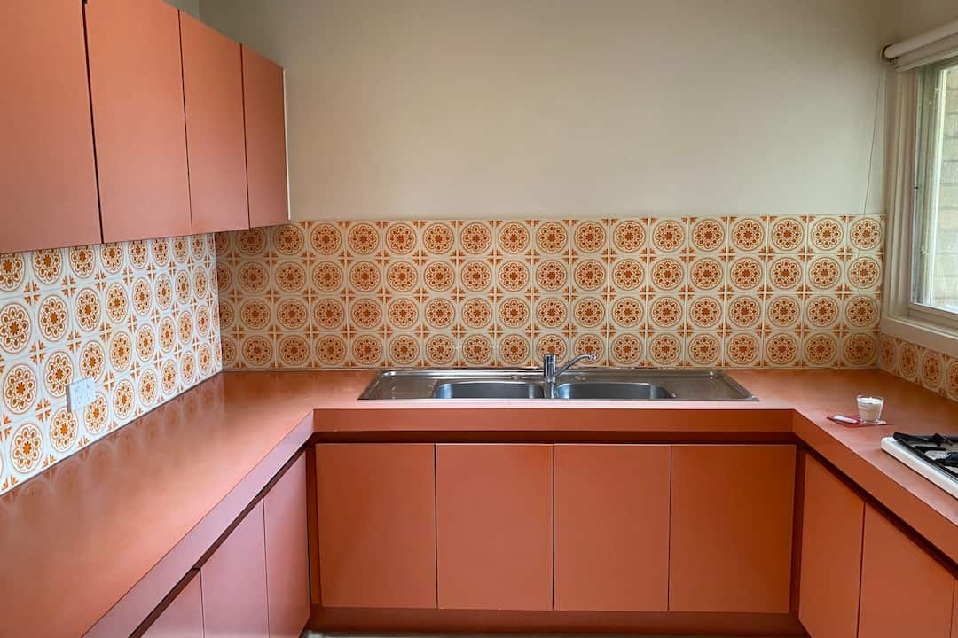 retro-style kitchen with orange laminate countertops and cabinets, patterned orange and cream tile backsplash, double stainless steel sink, window with natural light, minimal counter items
