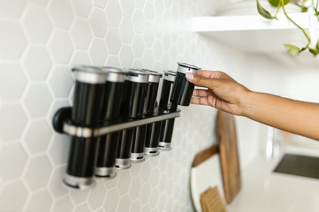 hand reaching for a black spice jar from a wall-mounted spice rack in a modern kitchen, white hexagon tile backsplash