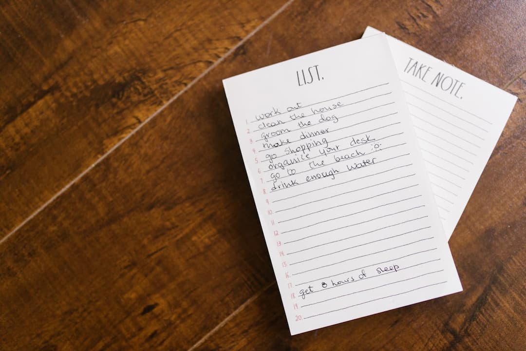 Handwritten to-do list on a white notepad placed on a wooden surface, listing daily tasks like working out, cleaning the house, and getting 8 hours of sleep