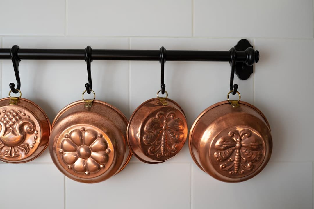 Four decorative copper pans with embossed designs, hanging on a black metal rod against a white tiled kitchen wall