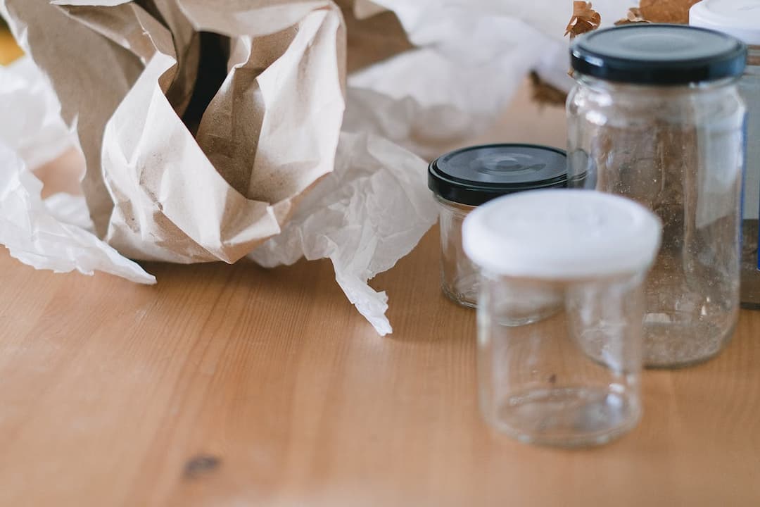 Empty glass jars with black and white lids, placed on a wooden table beside crumpled paper and tissue wrapping