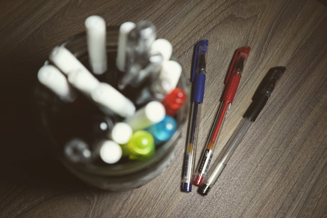 jar filled with assorted pens and markers, three ballpoint pens with red, blue, and black caps placed on wooden surface