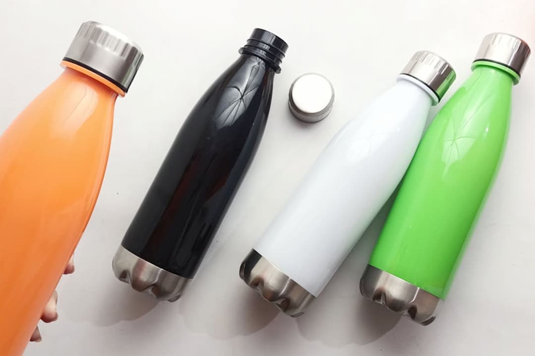 four stainless steel water bottles in orange, black, white, and green, silver caps, one bottle uncapped, placed on white surface with orange flowers in corner