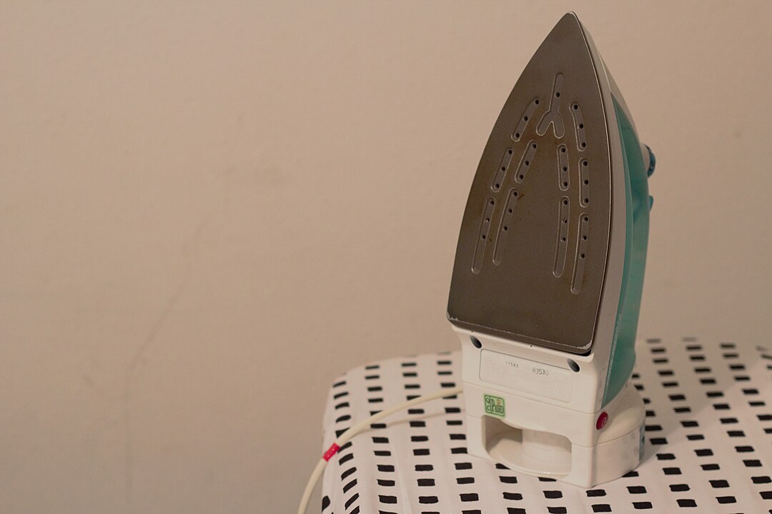 electric clothes iron, pointed soleplate with steam holes, resting upright on ironing board, white surface with black square pattern, power cord attached