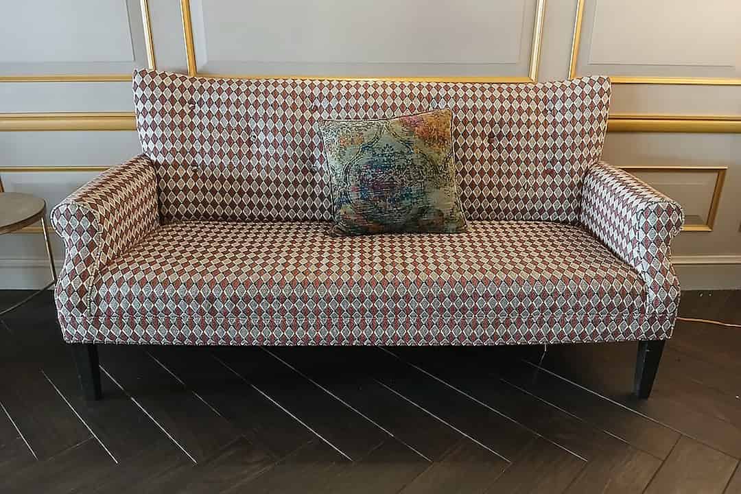 red and white diamond-patterned two-seater couch, single multicolored decorative pillow at center, black wooden legs, placed against a white and gold paneled wall, dark herringbone wooden floor