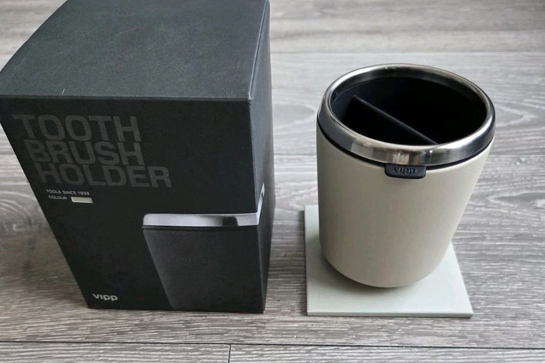 Vipp toothbrush holder with stainless steel rim, beige body, and black interior divider, placed beside its original black packaging on a wooden floor
