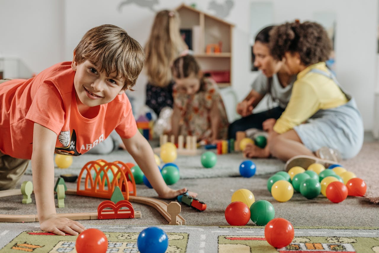 Smiling boy playing with toys on the floor while other children and an adult play in the background