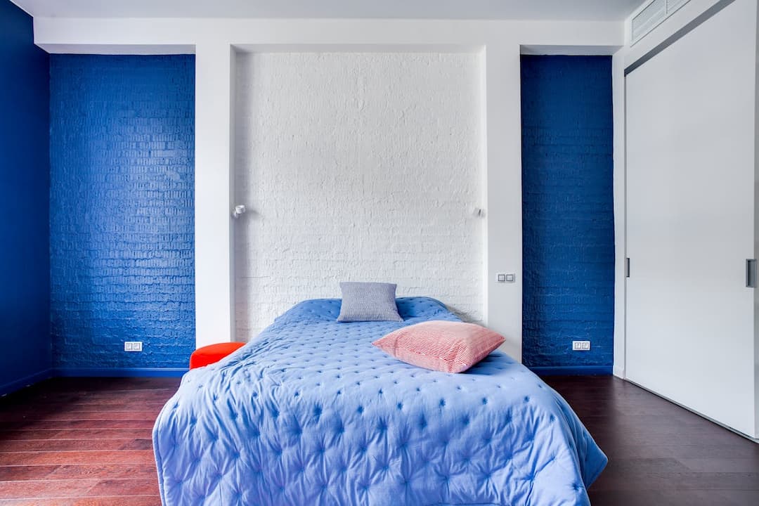 modern bedroom, blue tufted bedspread, red and grey throw pillows, white brick accent wall, deep blue side walls, dark wooden floor, sliding white closet door