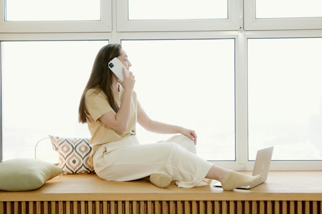 Woman sitting on a wooden window bench, talking on the phone, with a laptop and cushions beside her, large windows in the background