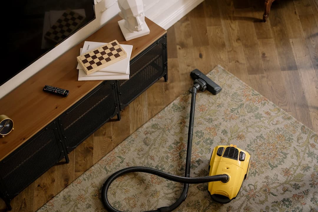 Yellow canister vacuum cleaner on a floral rug, next to a wooden TV console with books, a chessboard, a geometric bust, and a remote control