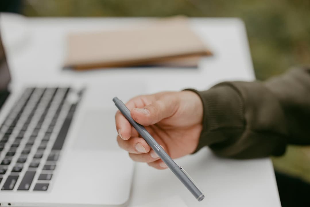 hand holding a gray pen, open laptop on white desk, brown notebooks in background